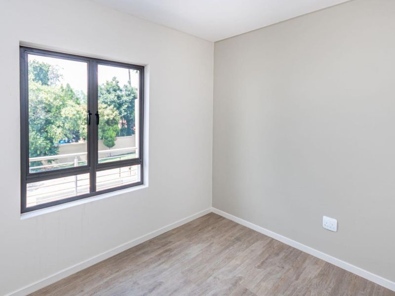 A bright, empty bedroom featuring modern laminate flooring, neutral wall tones, and a large aluminium-framed window providing natural light.