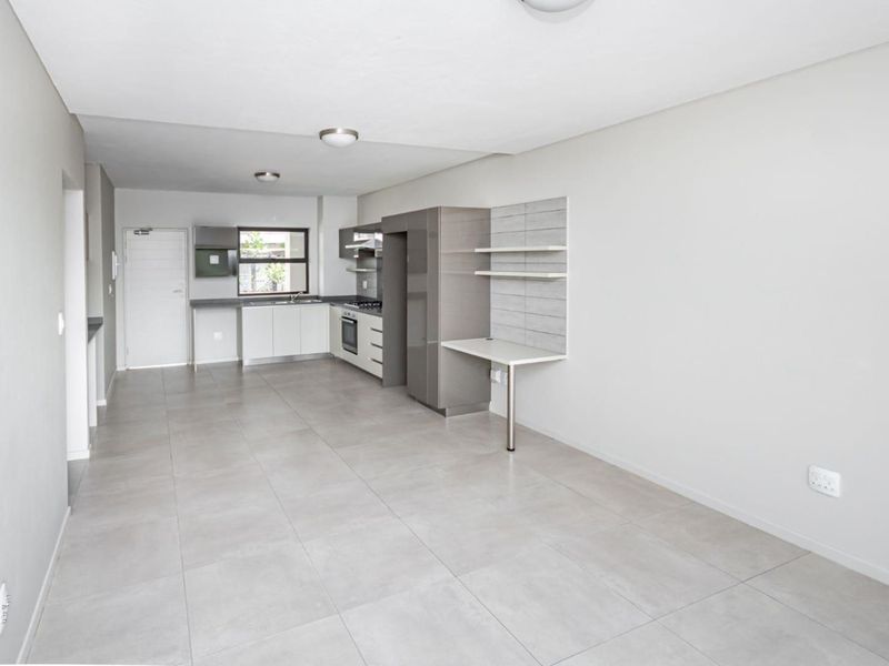 A bright, open-plan living and kitchen area featuring modern cabinetry, built-in appliances, a dedicated desk space, and neutral tiled flooring throughout.