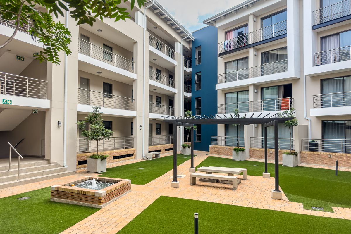 A well-maintained communal courtyard area featuring a pergola, brick-paved pathways, artificial lawn, and a small water feature surrounded by apartment building facades.