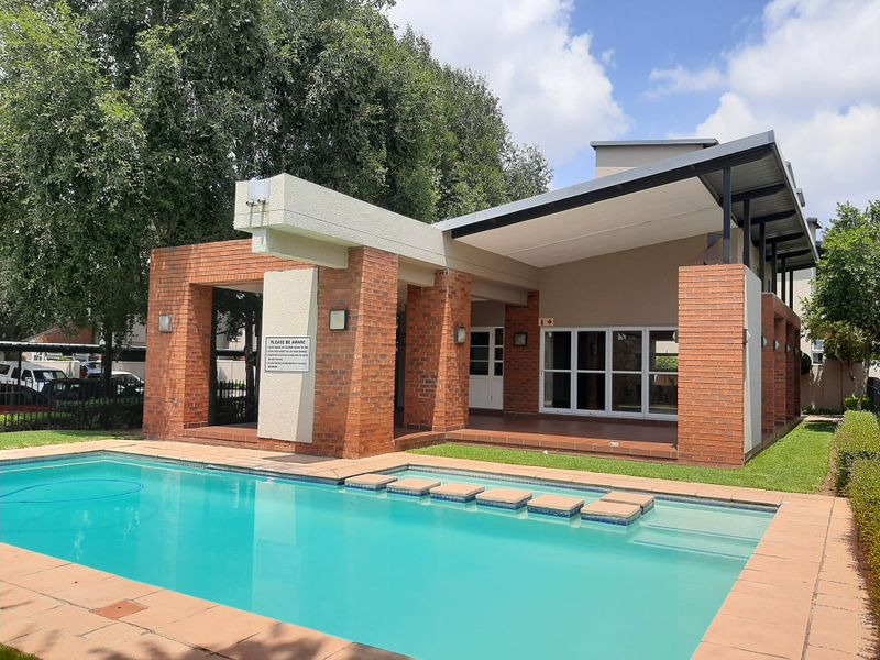 A well-maintained communal pool area featuring a brick-clad structure with a covered patio and paved surround, set within a landscaped garden environment.