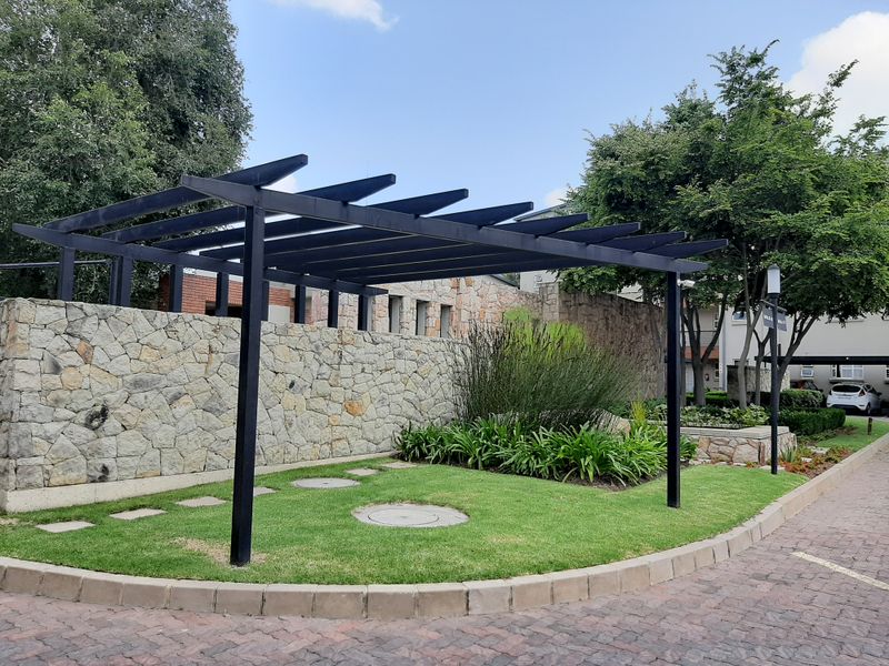 A well-maintained communal garden area featuring a black metal pergola, stone boundary wall, and manicured lawn within an apartment complex.