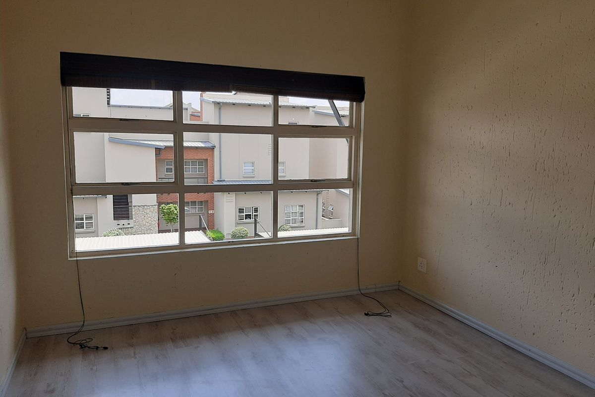 An empty bedroom featuring light-toned laminate flooring, a large aluminium-framed window with a roller blind, and neutral wall finishes.