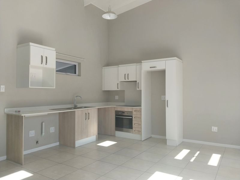 A clean, modern kitchen featuring light wood-tone cabinetry, white upper cabinets, integrated oven and hob, and neutral tiled flooring in an open-plan layout.