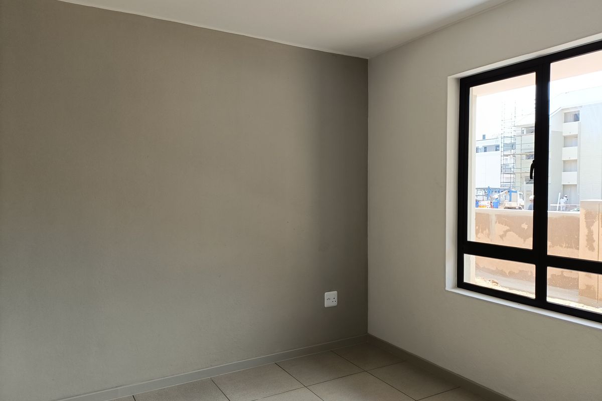 A bright, empty bedroom featuring neutral-toned walls, tiled flooring, and a large window with a dark frame.