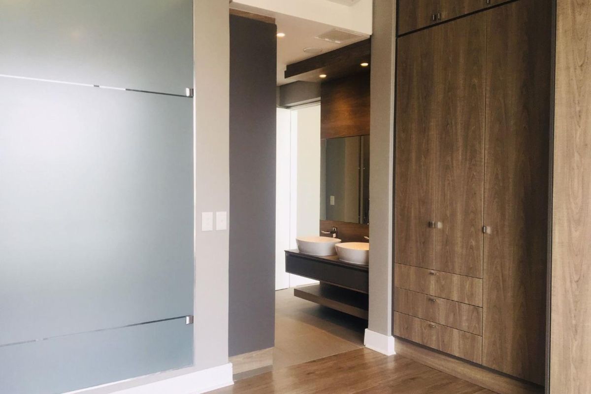 Modern bedroom featuring extensive built-in wooden cabinetry, laminate flooring, and a sleek frosted glass door leading to an en-suite bathroom with dual vanity.