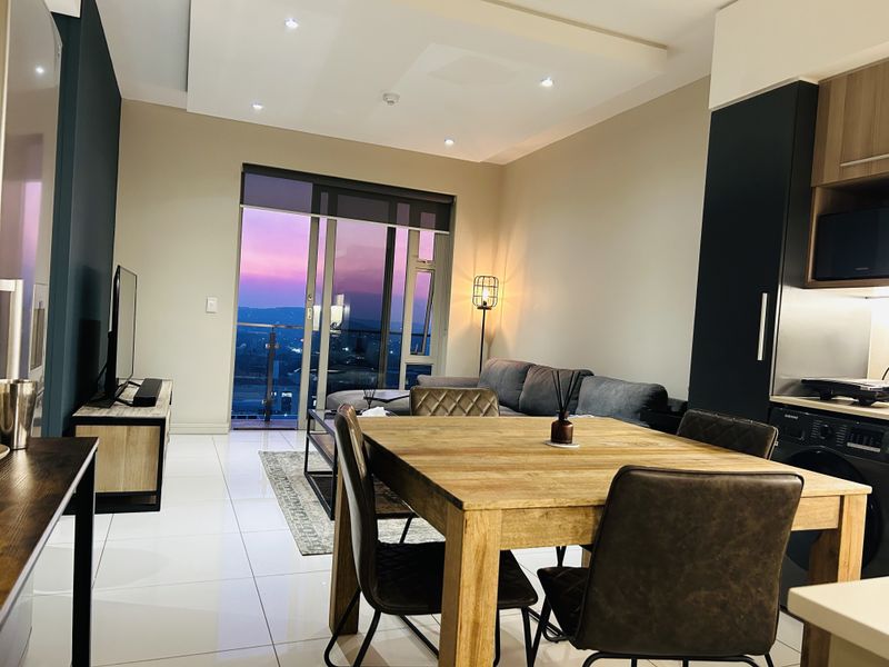 Modern open-plan living and dining area featuring sleek tiled floors, recessed lighting, and a sliding door leading to a balcony with sunset views.