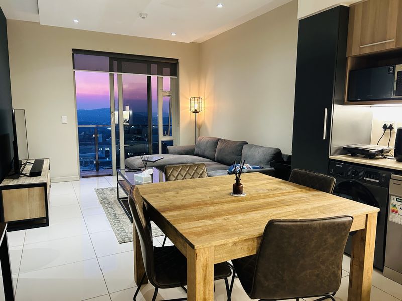 Modern open-plan apartment living area featuring a sleek kitchen, tiled floors, and a sliding door leading to a balcony with sunset views.