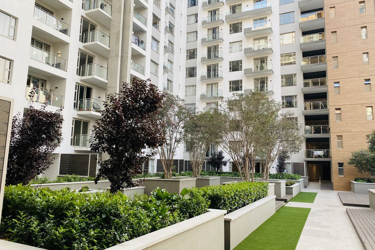 A well-maintained apartment complex courtyard featuring landscaped planters, artificial turf, paved walkways, and multiple levels of balconies with glass balustrades.