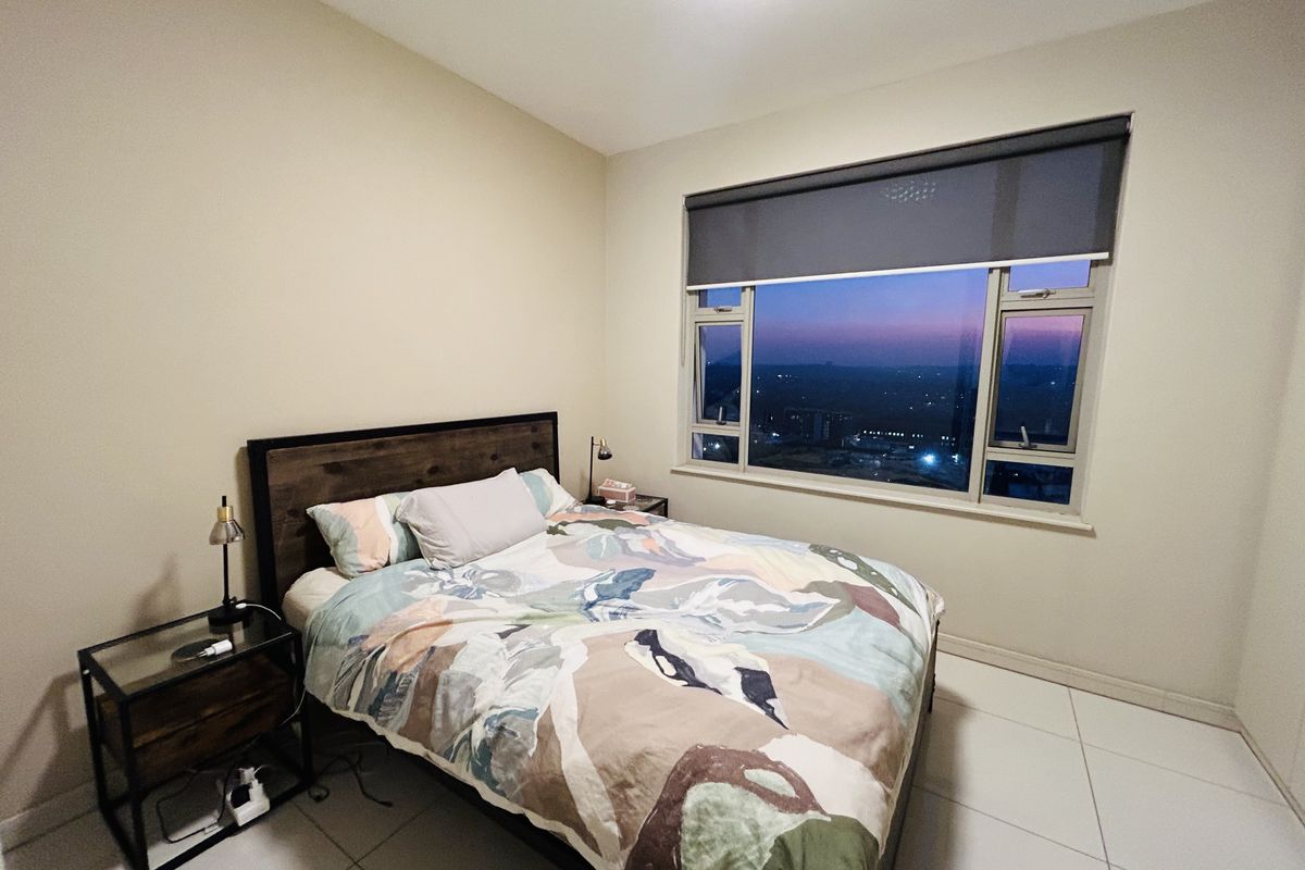 A well-lit bedroom featuring large windows with roller blinds, tiled flooring, and a pleasant evening view.