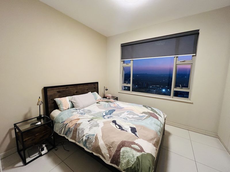 A well-lit bedroom featuring large windows with roller blinds, tiled flooring, and a pleasant evening view.