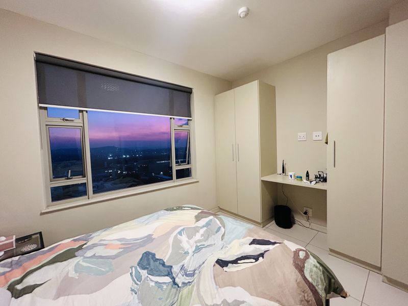 A well-maintained bedroom featuring built-in wardrobes, a integrated desk area, tiled flooring, and a large window offering scenic sunset views.