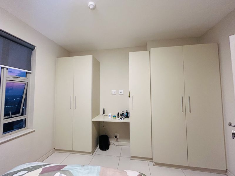 A bedroom featuring neutral-toned built-in cupboards with an integrated desk space, tiled flooring, and a window with a roller blind.