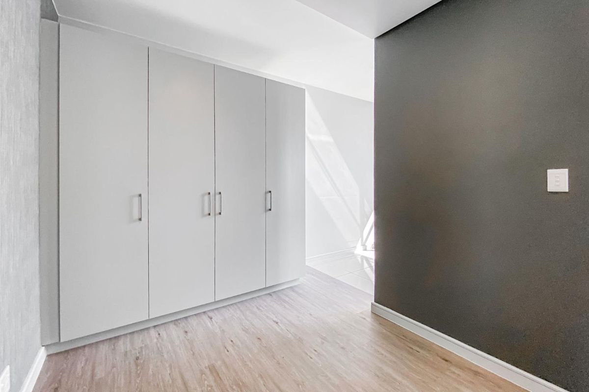 A bright, modern bedroom featuring neutral laminate flooring, clean white built-in cupboards, and a contrasting dark accent wall.