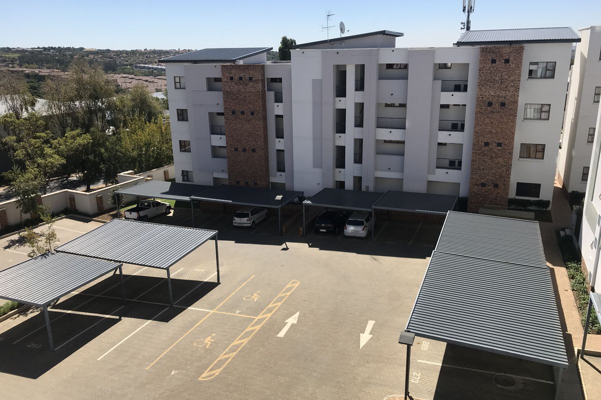 Aerial view of the apartment complex showing the building facade, paved parking area, and multiple covered parking bays.