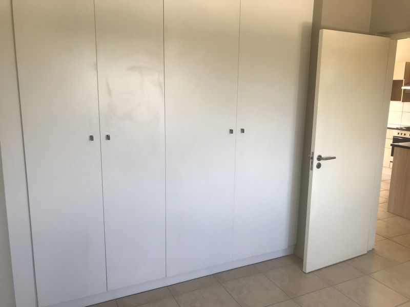 A clean, empty bedroom featuring neutral tiled flooring and ample built-in cupboard storage, with a view into the adjacent kitchen area.