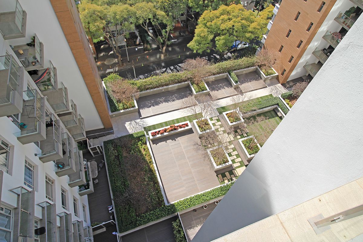 An elevated aerial view of the apartment complex courtyard, featuring landscaped garden beds, paved walkways, and wooden decking areas between the building wings.