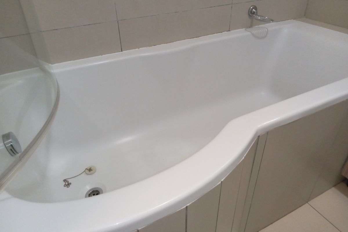 A clean, modern bathroom featuring a white bathtub with a glass shower screen and neutral wall tiling.