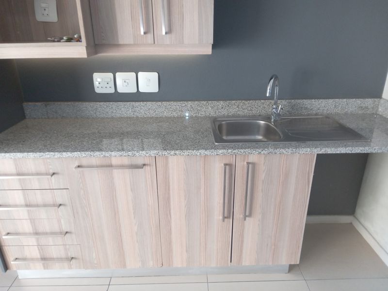 A modern kitchen area featuring light wood-grain cabinetry, granite countertops, a stainless steel sink, and tiled flooring in a clean, well-maintained condition.