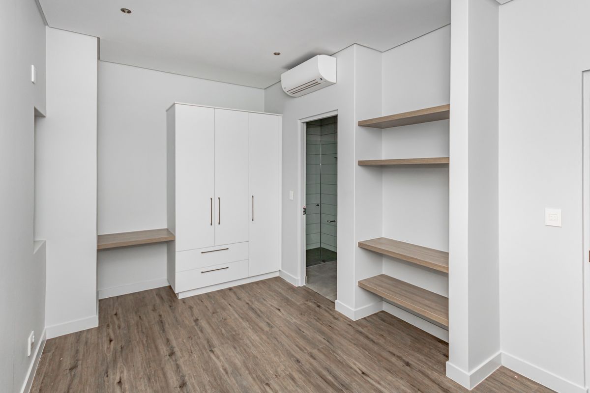 A modern, empty bedroom featuring built-in wardrobes, integrated shelving, a built-in desk, air conditioning, and wood-look laminate flooring.