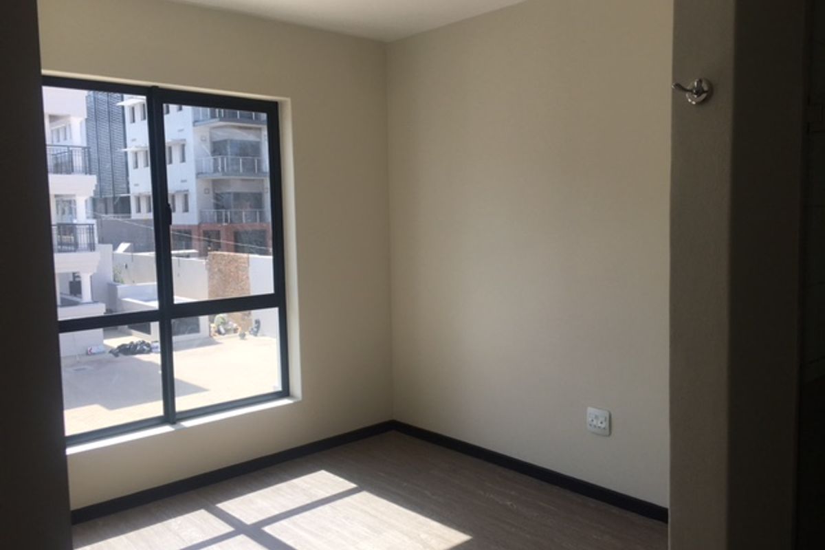 A bright, empty bedroom featuring modern laminate flooring, neutral walls, and a large window with views of the surrounding apartment complex.
