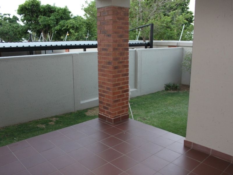 A covered patio area featuring tiled flooring and a brick support pillar, overlooking a private garden space with a concrete boundary wall.