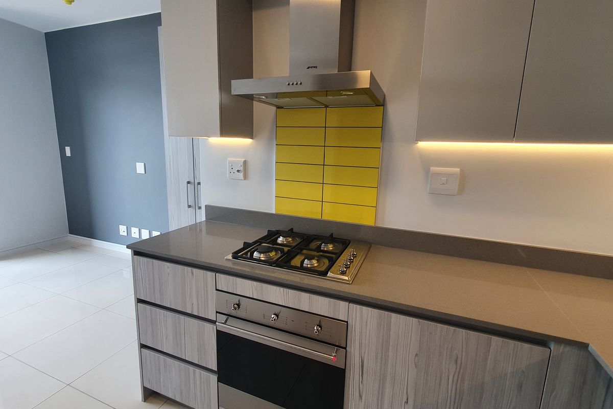 Modern kitchen featuring sleek grey cabinetry, stone countertops, a built-in gas hob, oven, and a vibrant yellow tiled backsplash with integrated under-cabinet lighting.