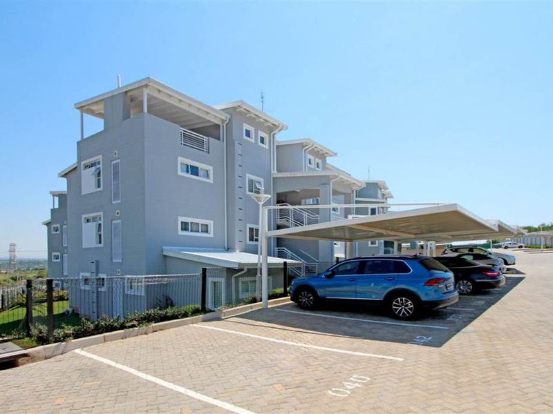Exterior view of a modern multi-story apartment complex featuring paved parking bays with covered carports and perimeter fencing.