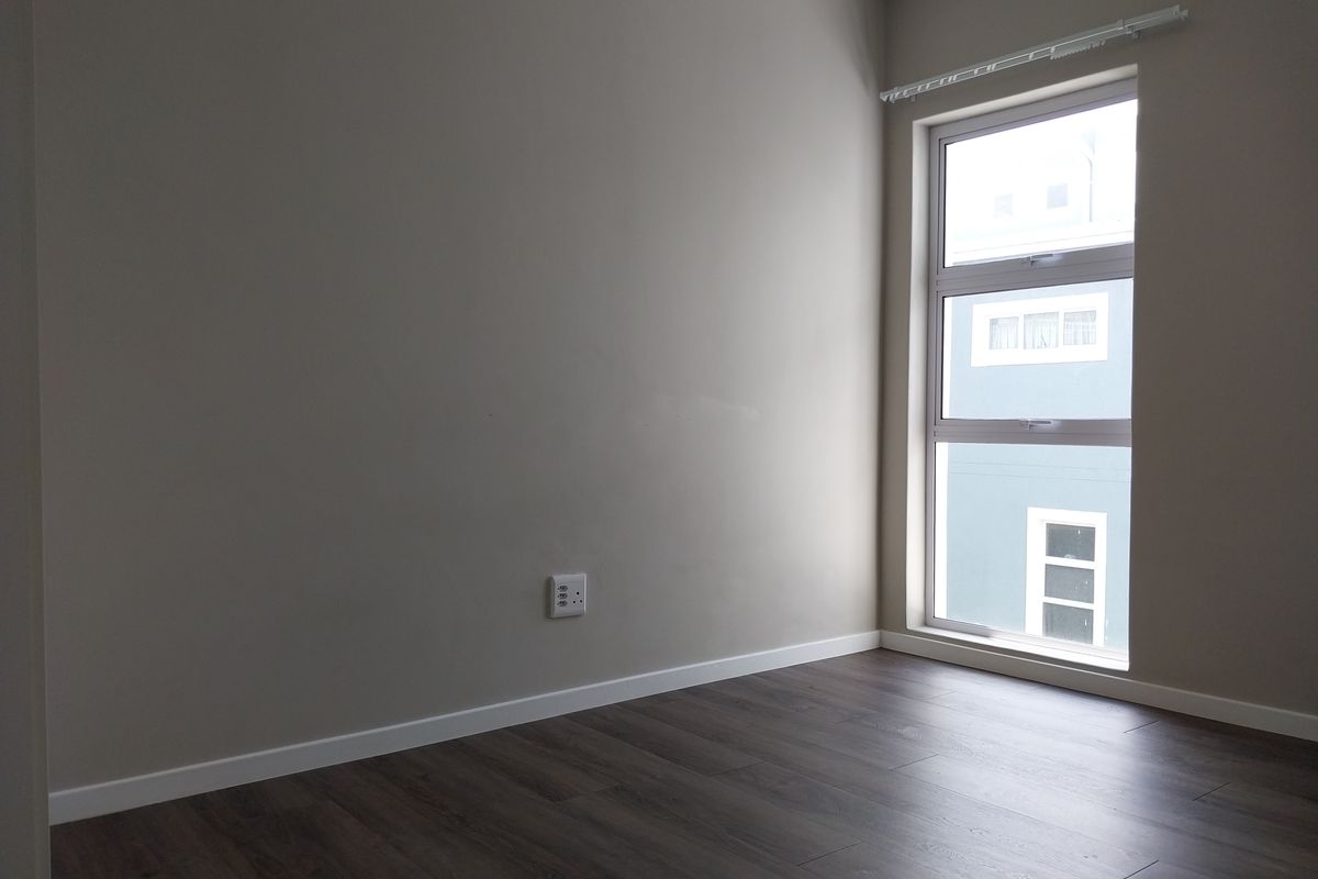 A clean, empty bedroom featuring modern dark laminate flooring, neutral wall paint, and a large aluminium-framed window providing natural light.