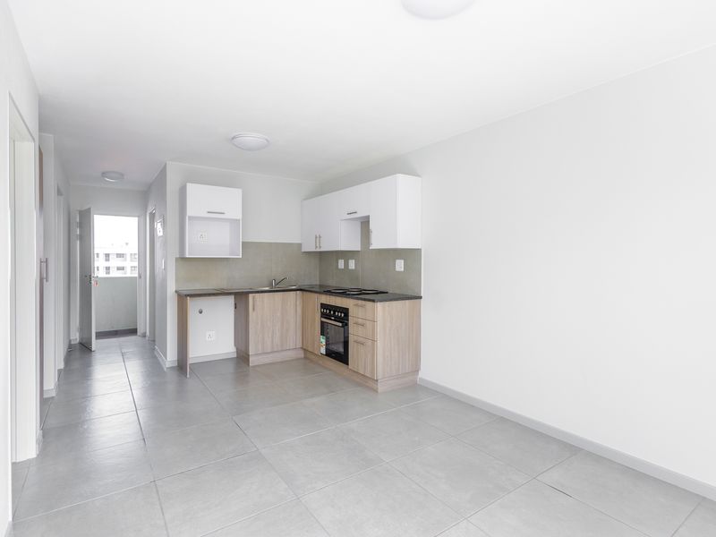 Modern open-plan kitchen featuring light wood-finish cabinetry, dark countertops, built-in oven and hob, and neutral tiled flooring in a bright, vacant apartment space.