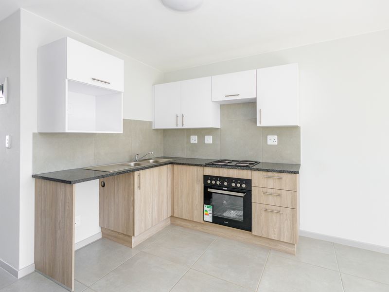 A clean, modern kitchen featuring light wood-finish cabinetry, dark countertops, a built-in oven and hob, and neutral tiled flooring.