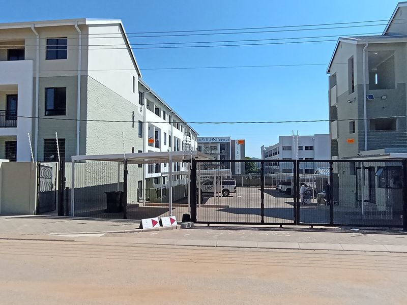 Street view of Brooke Manor apartment complex entrance featuring secure gated access, perimeter wall with electric fencing, and multi-story residential buildings.