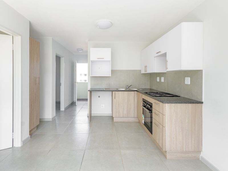 A clean, modern kitchen featuring light wood-finish cabinetry, granite-look countertops, built-in oven and hob, and neutral tiled flooring in an open-plan layout.