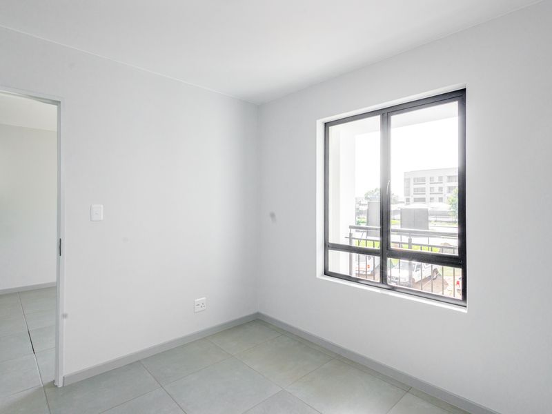 A bright, empty bedroom featuring neutral tiled flooring, clean white walls, and a large aluminium-framed window allowing for natural light.