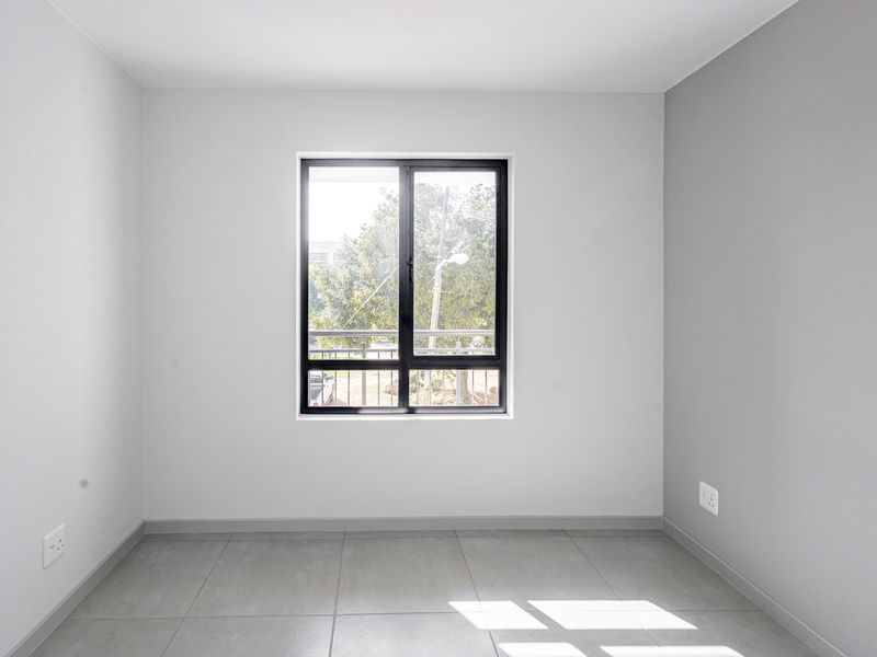 A bright, empty bedroom featuring neutral grey walls, tiled flooring, and a modern black aluminium window frame.