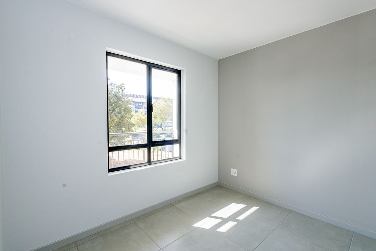A bright, empty bedroom featuring neutral wall tones, tiled flooring, and a large aluminium-framed window providing natural light.