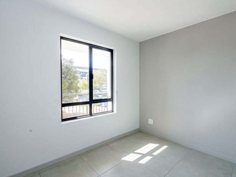 A bright, empty bedroom featuring neutral wall tones, tiled flooring, and a large aluminium-framed window providing natural light.