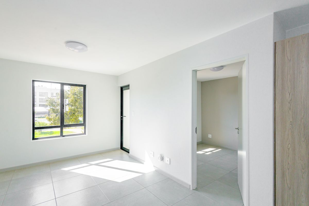 A bright, modern, and empty living area featuring neutral tiled flooring, clean white walls, and large aluminium-framed windows allowing for ample natural light.