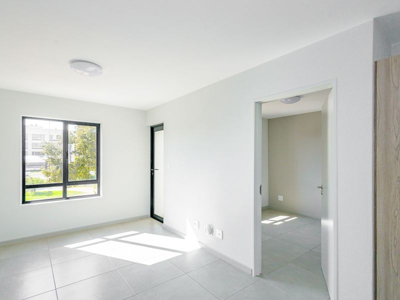 A bright, modern, and empty living area featuring neutral tiled flooring, clean white walls, and large aluminium-framed windows allowing for ample natural light.