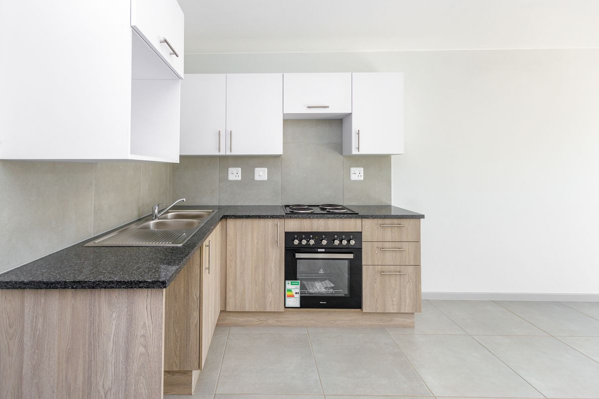 A modern, clean kitchen featuring light wood-finish cabinetry, dark granite-look countertops, a built-in oven and hob, and neutral tiled flooring.