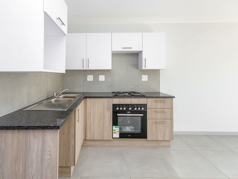 A modern, clean kitchen featuring light wood-finish cabinetry, dark granite-look countertops, a built-in oven and hob, and neutral tiled flooring.