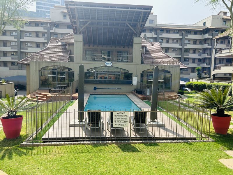 A well-maintained communal swimming pool area featuring secure metal fencing, paved surrounds, and landscaped garden space within the apartment complex.