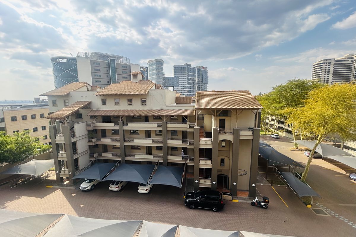 An elevated aerial view of a multi-story apartment complex featuring a tiled roof, paved parking area with shade sails, and surrounding urban landscape.