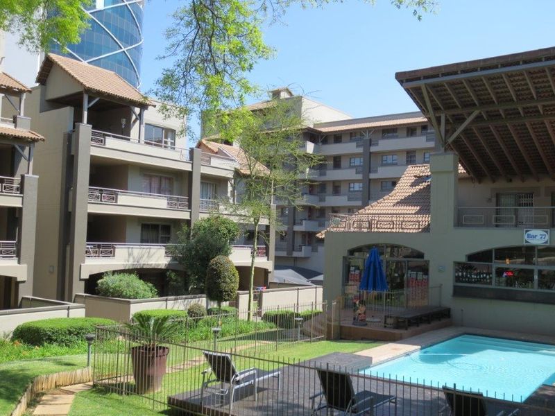 Communal apartment complex pool area featuring a swimming pool, landscaped gardens, and covered patio space with surrounding multi-story residential buildings.