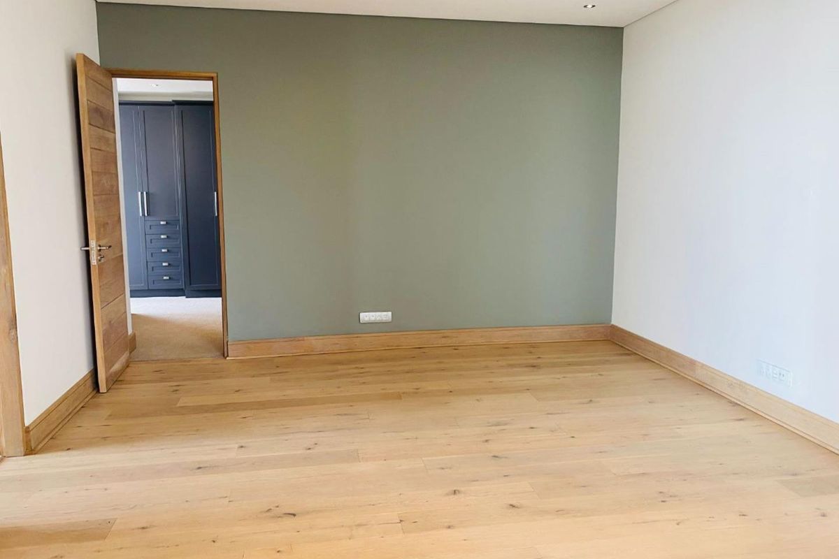 A bright, empty bedroom featuring light oak timber flooring, a sage green feature wall, and recessed ceiling lighting, with an adjacent walk-in closet.