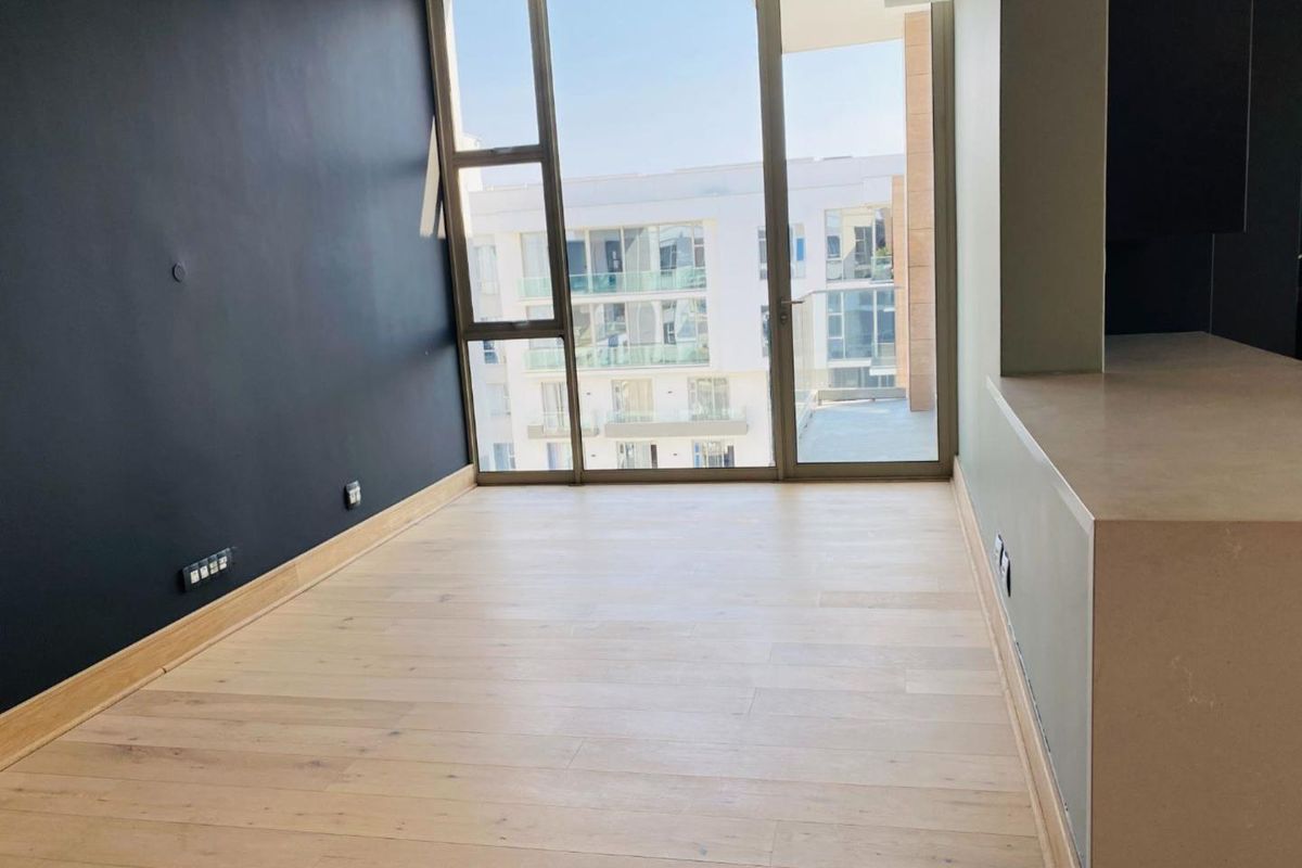 A bright, empty living area featuring light wood flooring, a dark accent wall, and floor-to-ceiling windows providing access to a balcony.