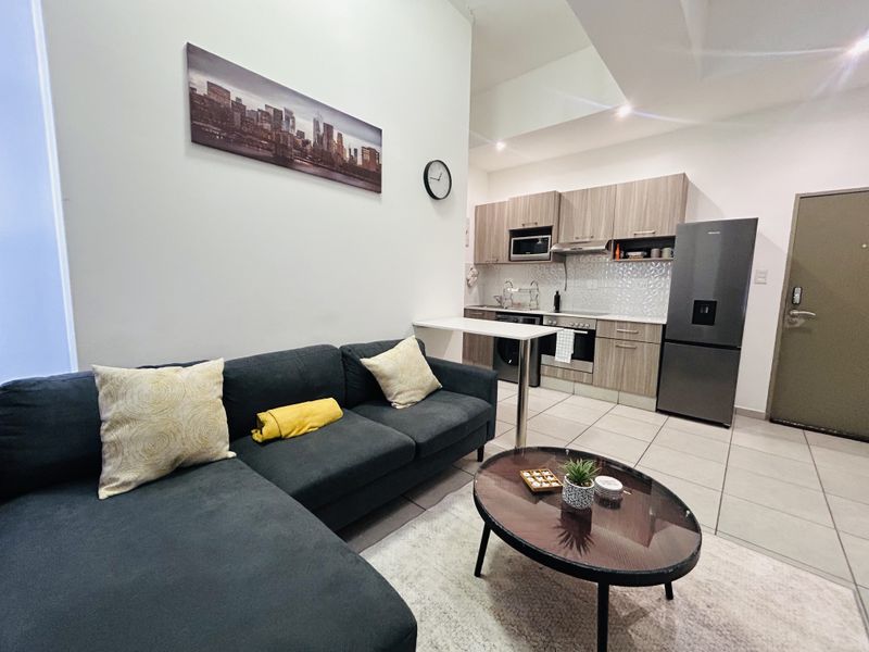 Modern open-plan living area featuring a compact kitchen with built-in cabinetry, integrated appliances, and a breakfast bar, seamlessly connected to the lounge space.