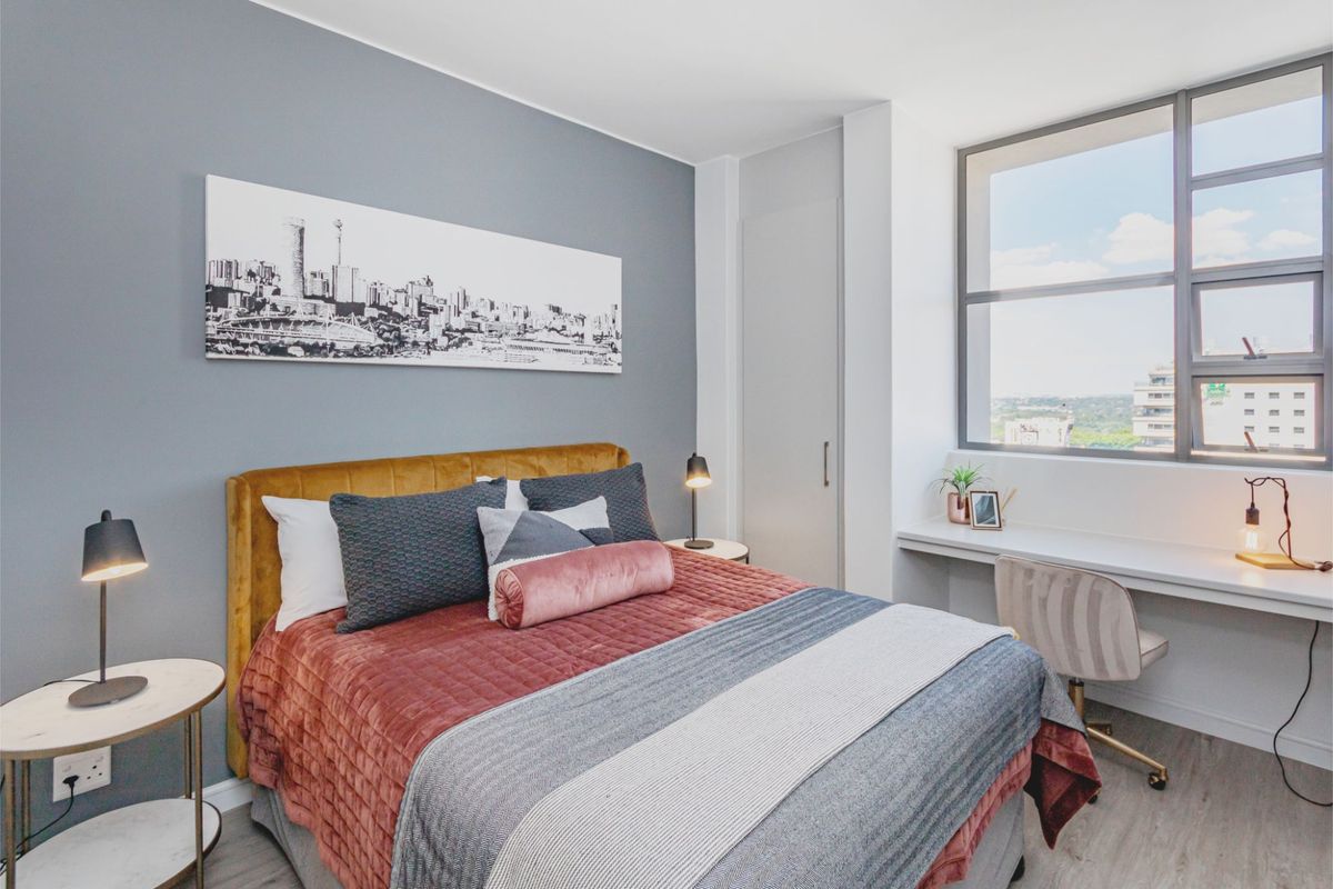 A well-lit bedroom featuring a built-in desk, integrated wardrobe, and large window offering natural light and urban views.