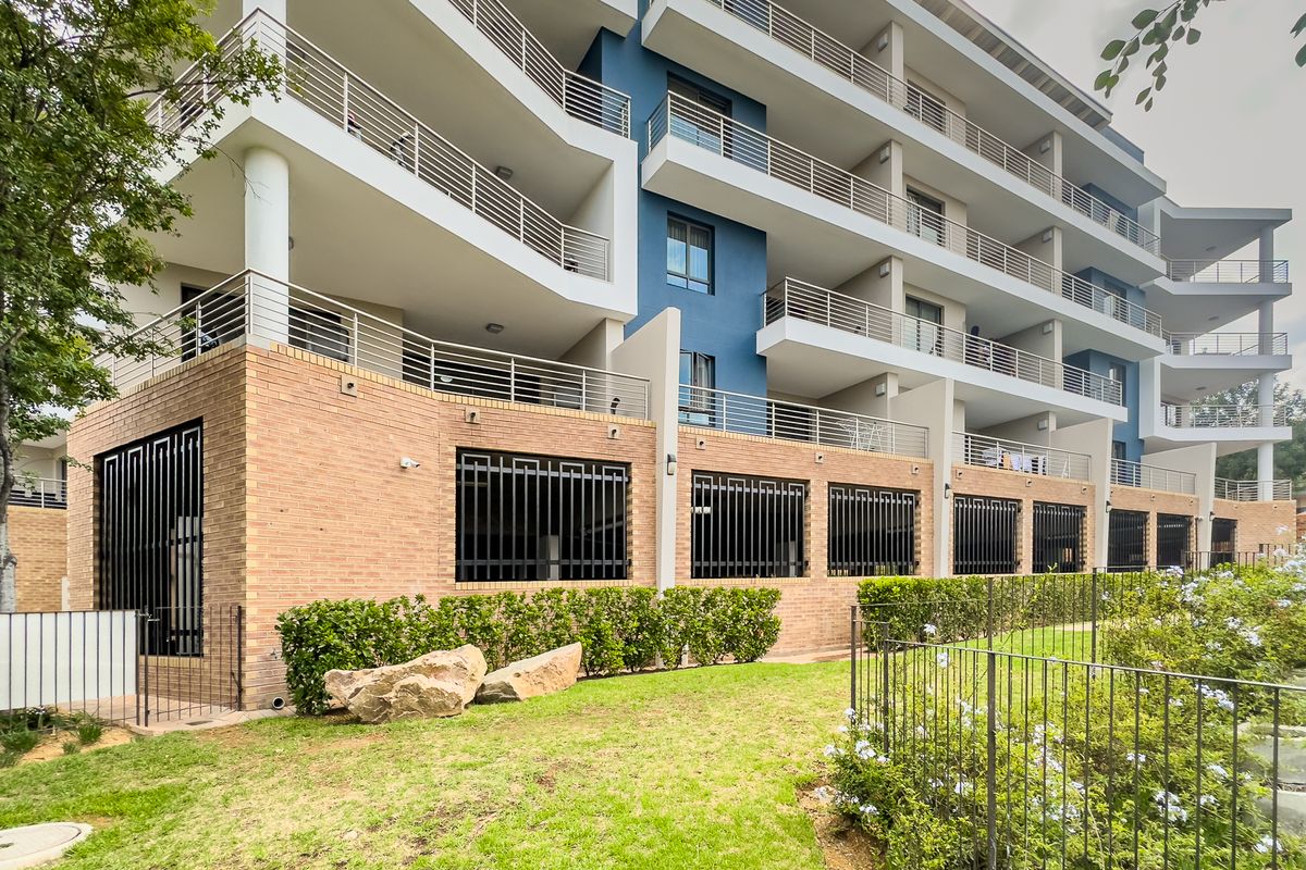 Rear view of a modern apartment complex featuring multi-level balconies, brick lower facade with security grilles, and a well-maintained landscaped garden area.