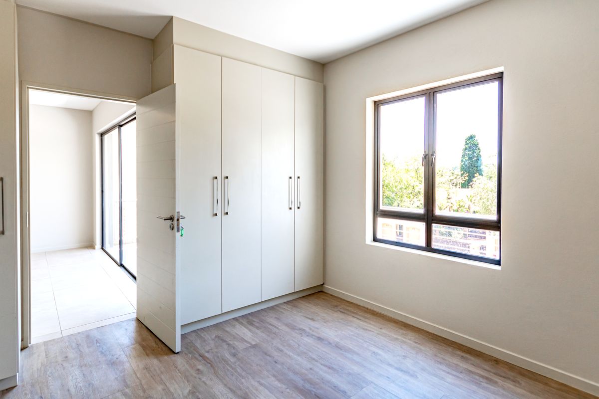 A bright, empty bedroom featuring modern laminate flooring, built-in cupboards, and a large window providing natural light.