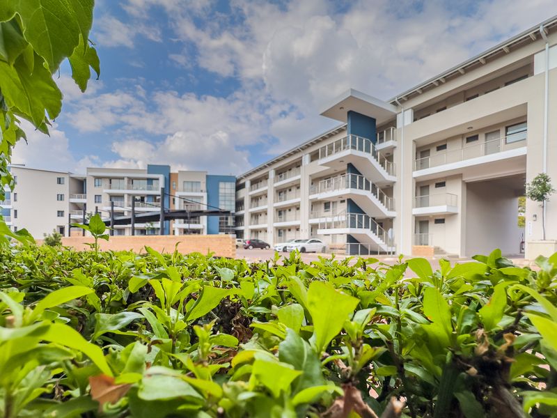 A view of a modern multi-story apartment complex featuring external staircases, balconies, and a paved driveway, framed by foreground garden hedging.